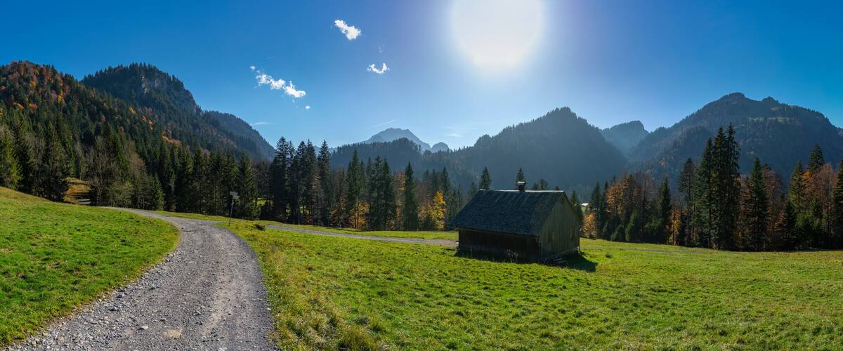 A rural landscape with a dirt road, a small wooden cabin, and a mountain range under a clear blue sky.