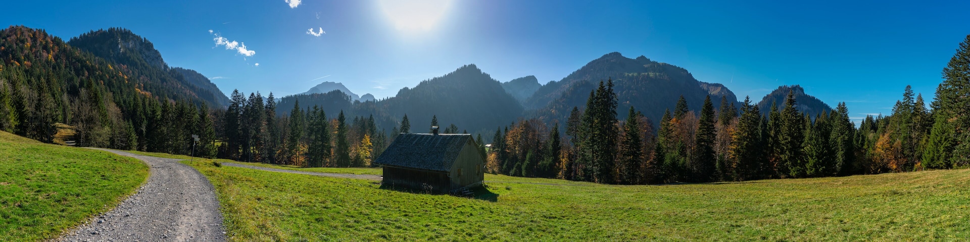 A rural landscape with a dirt road, a small wooden cabin, and a mountain range under a clear blue sky.