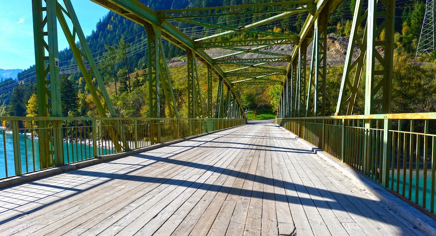 Uma ponte de madeira com grades de metal, cercada por montanhas arborizadas e um corpo de água.
