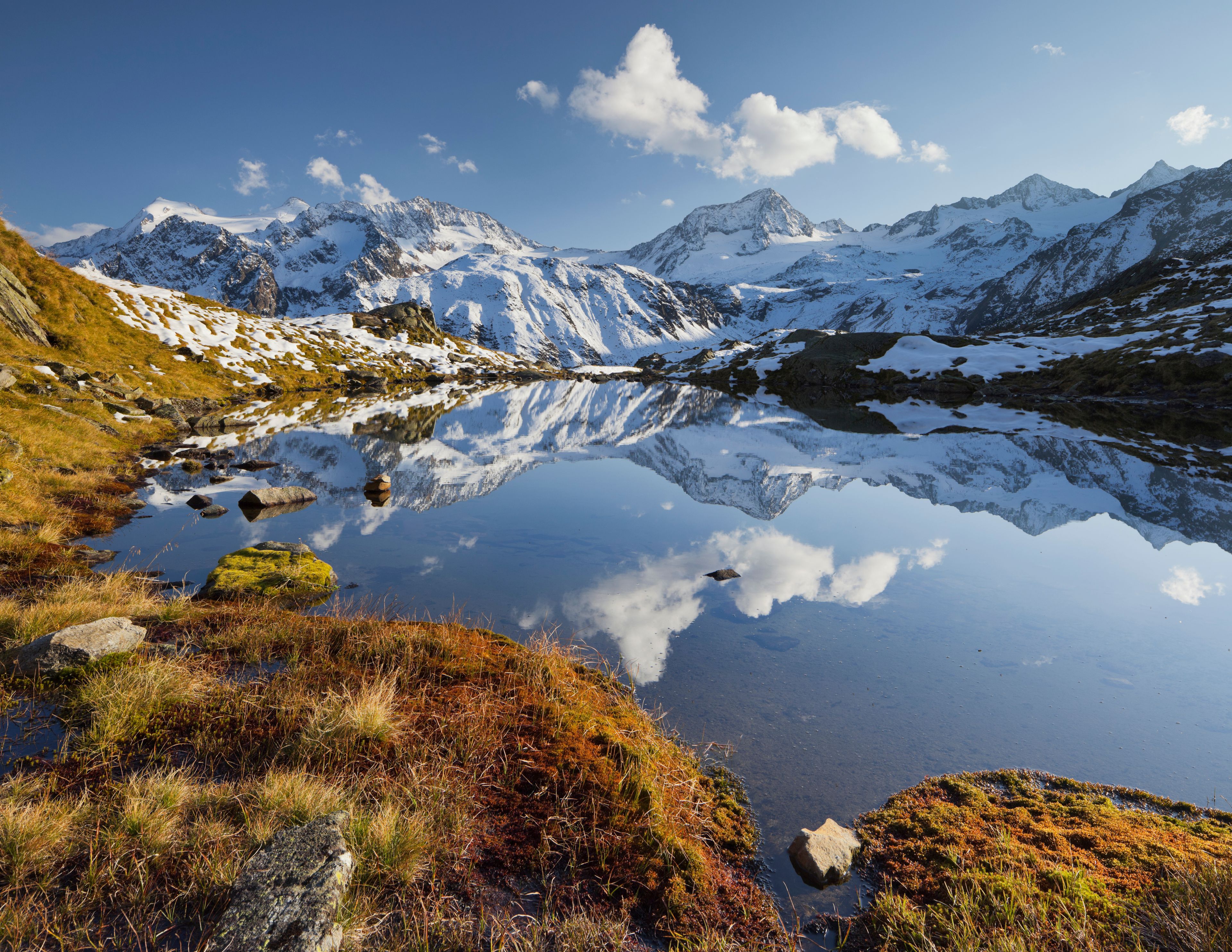 A mountain lake with reflections of the peaks, surrounded by snow-covered mountains and patches of grass.