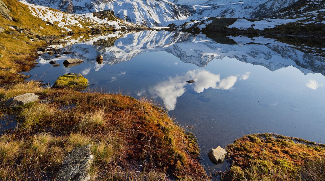A mountain lake with reflections of the peaks, surrounded by snow-covered mountains and patches of grass.