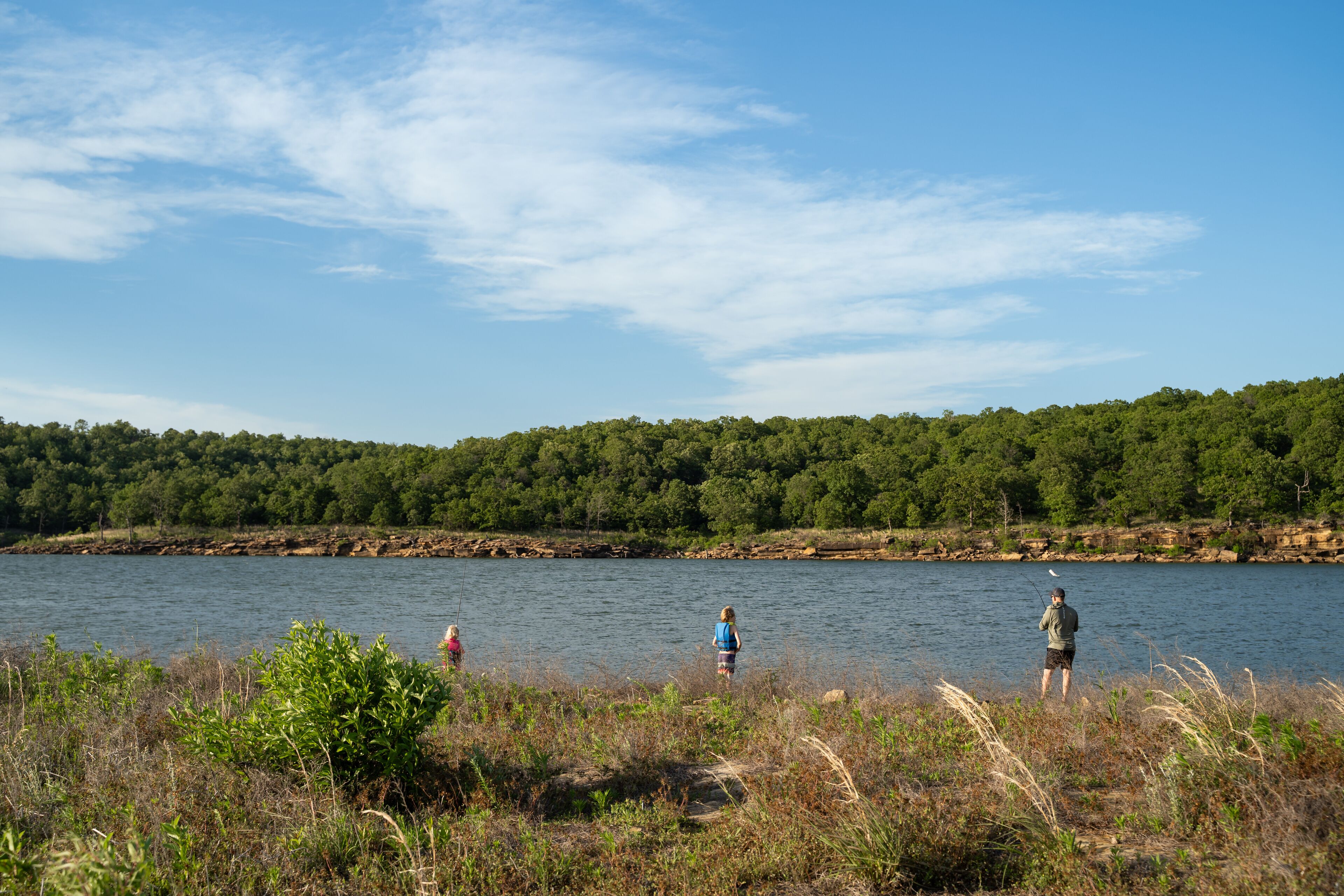 A calm lake surrounded by dense forest, with people fishing and a child playing nearby.