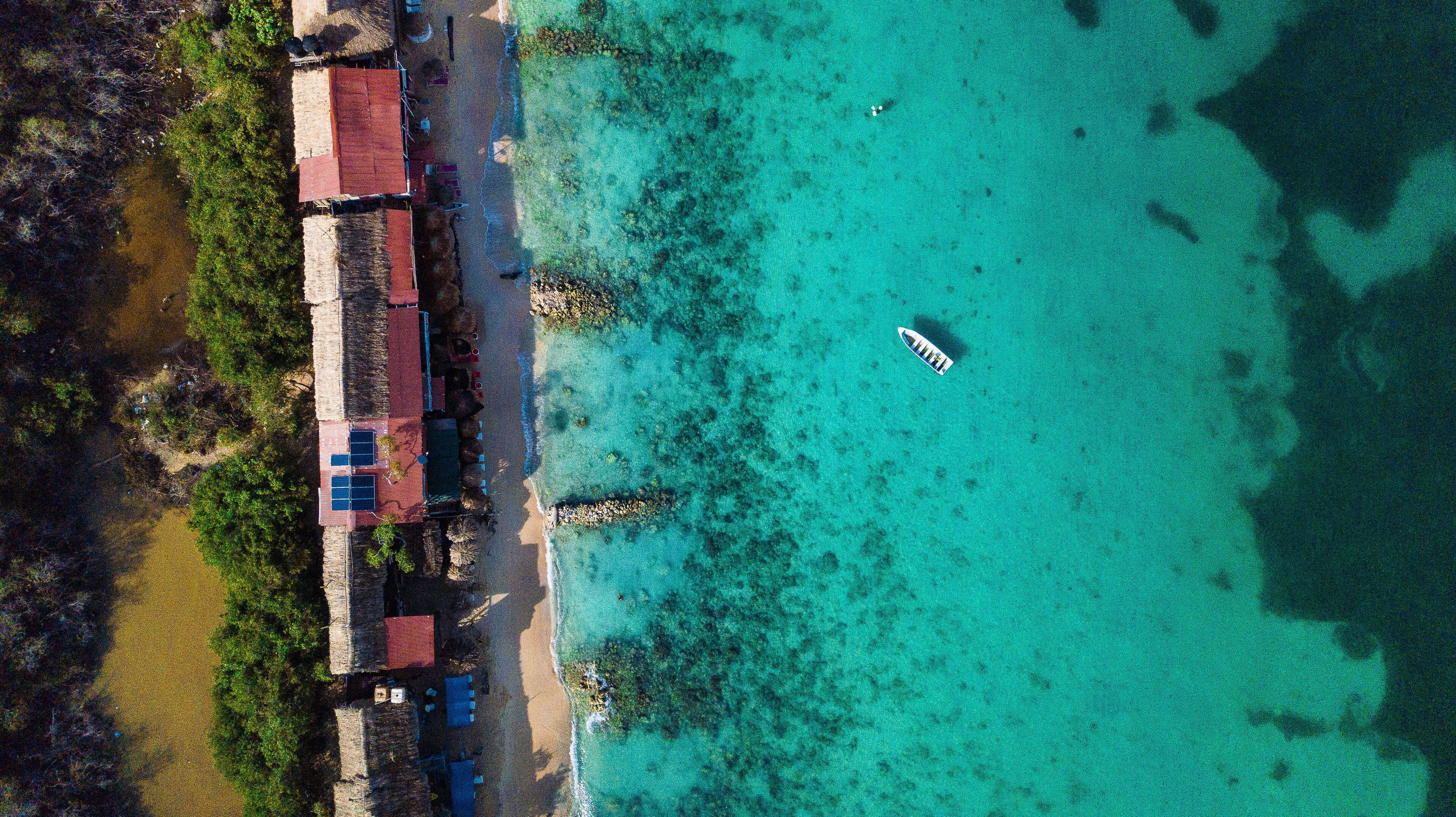 An aerial view of a coastal area with clear turquoise waters, a small boat, and buildings with red roofs near the shore.