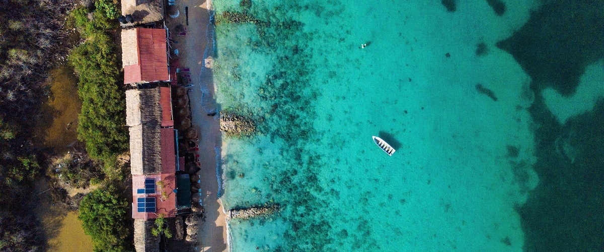 An aerial view of a coastal area with clear turquoise waters, a small boat, and buildings with red roofs near the shore.