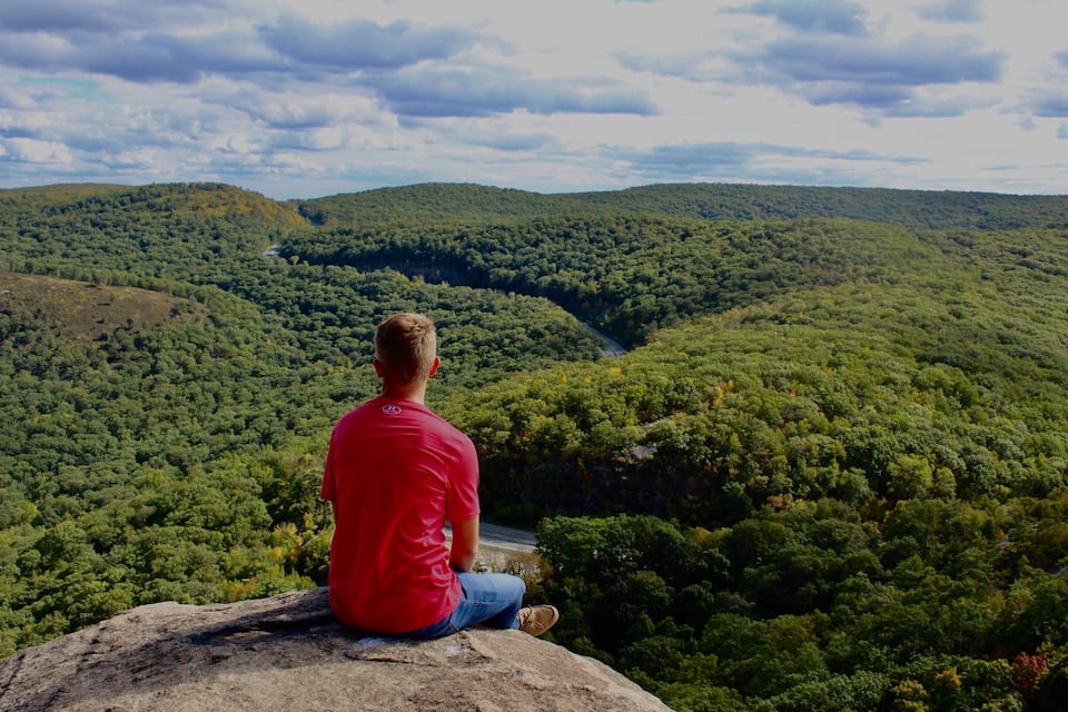 A man in a red shirt sitting on a rock overlooking a dense forested valley.