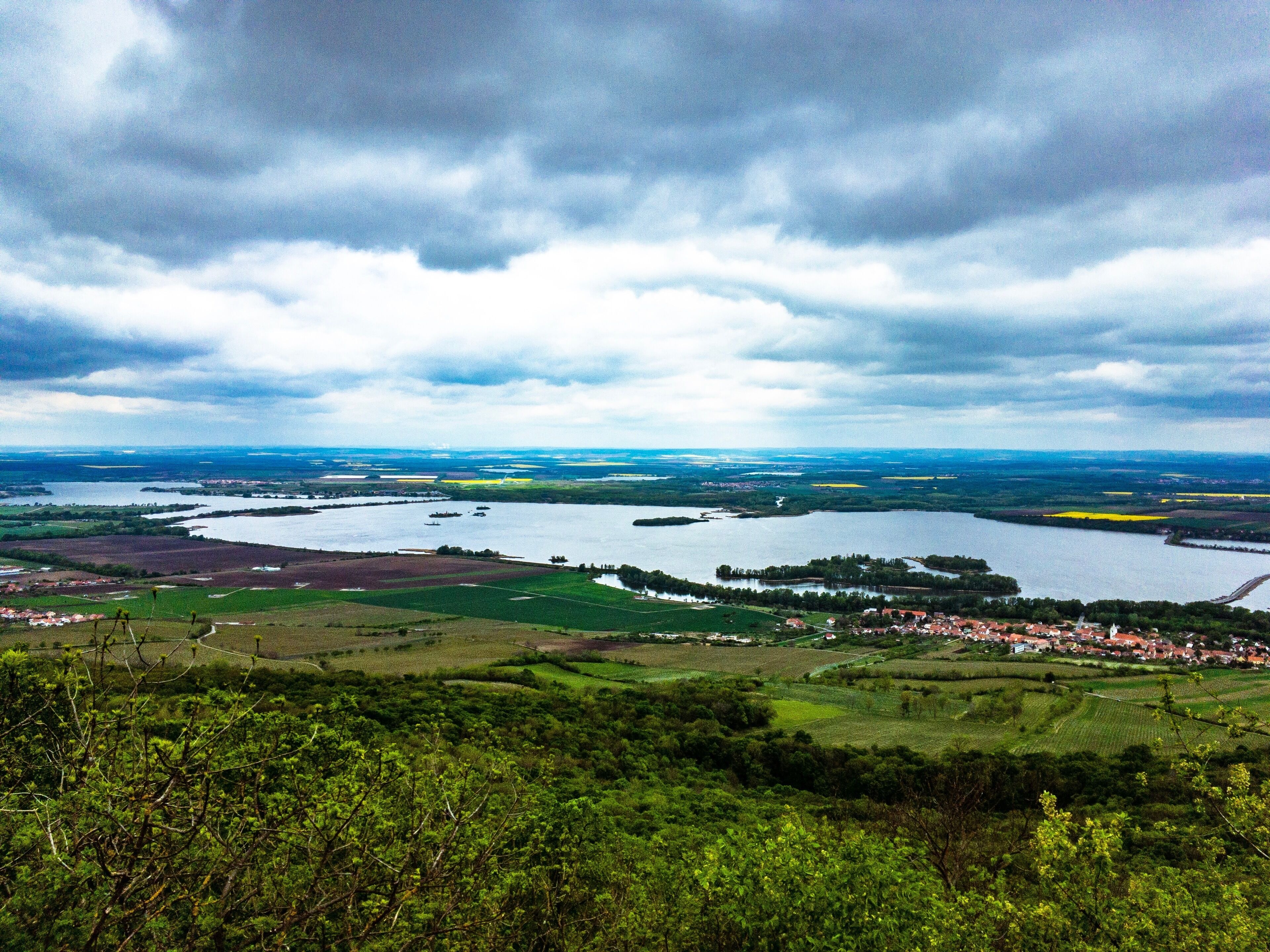 A panoramic view of a river valley with fields, a small town, and a cloudy sky.
