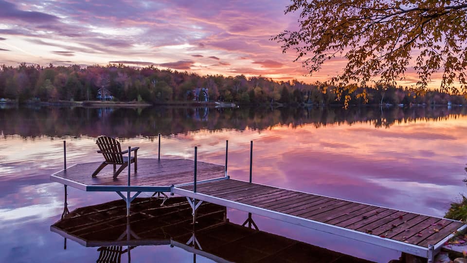 A wooden dock extending into a calm lake at sunset with a chair on the left.