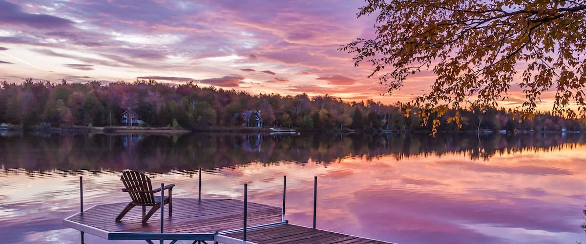 A wooden dock extending into a calm lake at sunset with a chair on the left.