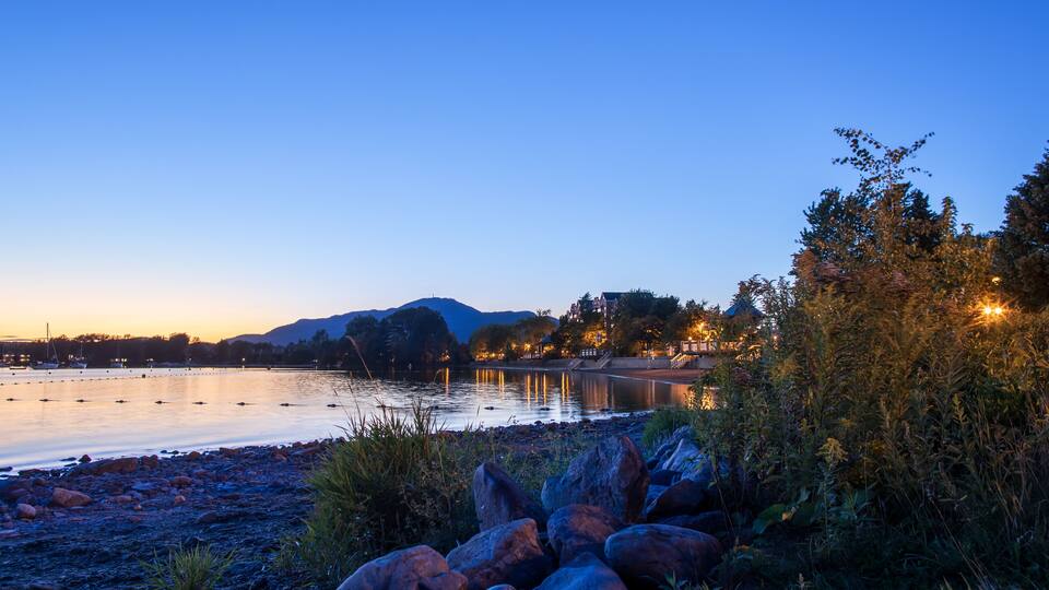 A calm lake at dusk with rocks and vegetation in the foreground, a distant shoreline with buildings, and mountains in the background.