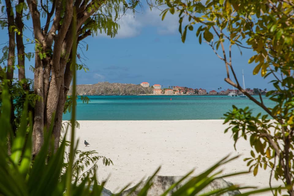 A beach with turquoise water, white sand, and buildings in the background.