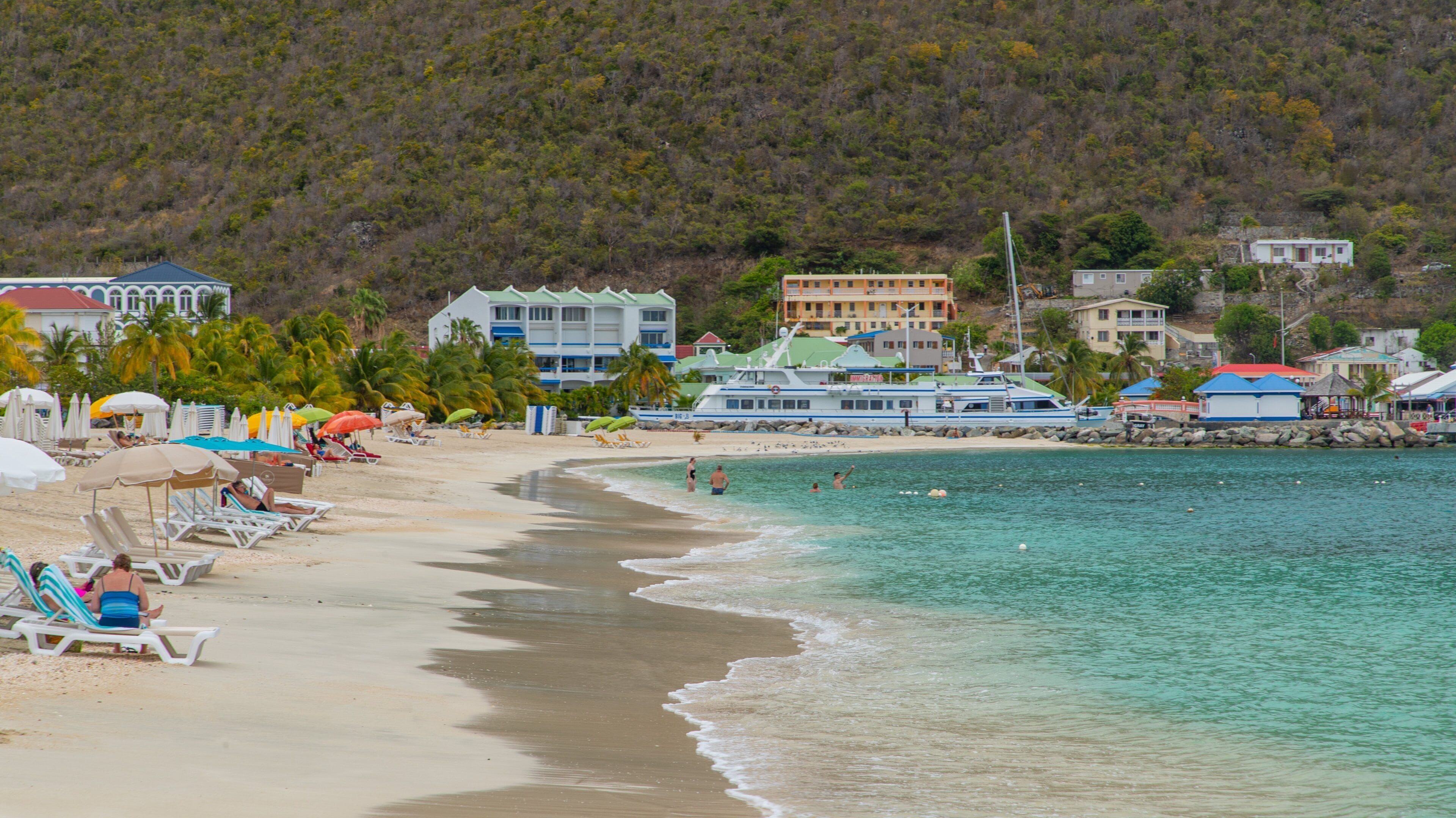 A beach with clear turquoise water, white sandy shore, and colorful umbrellas. There are buildings and a boat in the background.