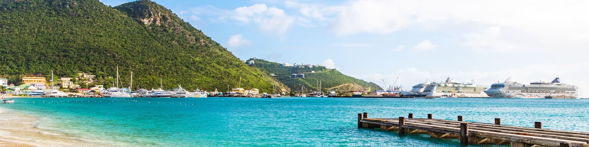 Una playa turquesa con un muelle de madera, una montaña al fondo y cruceros anclados en el agua.