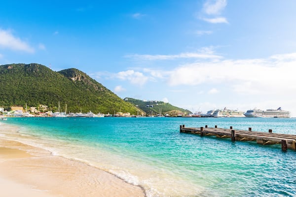 Uma praia turquesa com um píer de madeira, uma montanha ao fundo e navios de cruzeiro atracados na água.