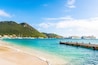 A turquoise beach with a wooden pier, a mountain in the background, and cruise ships docked in the water.