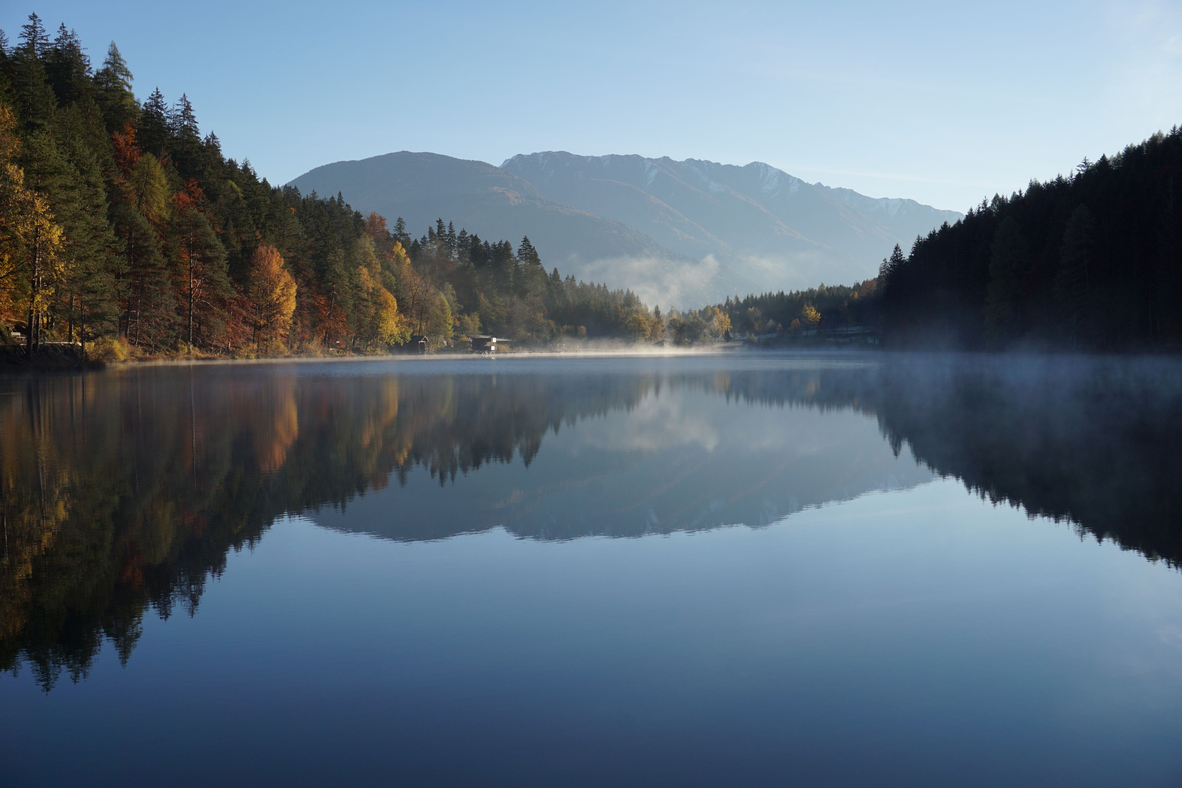 Ein ruhiger See mit der Spiegelung von Bäumen und Bergen im Hintergrund.