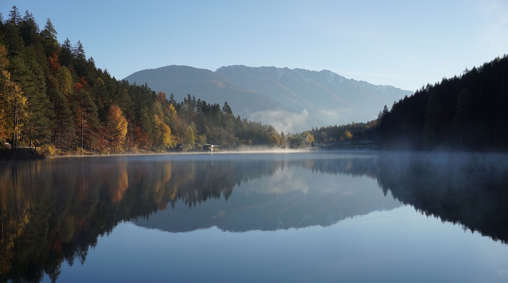 Ein ruhiger See mit der Spiegelung von Bäumen und Bergen im Hintergrund.