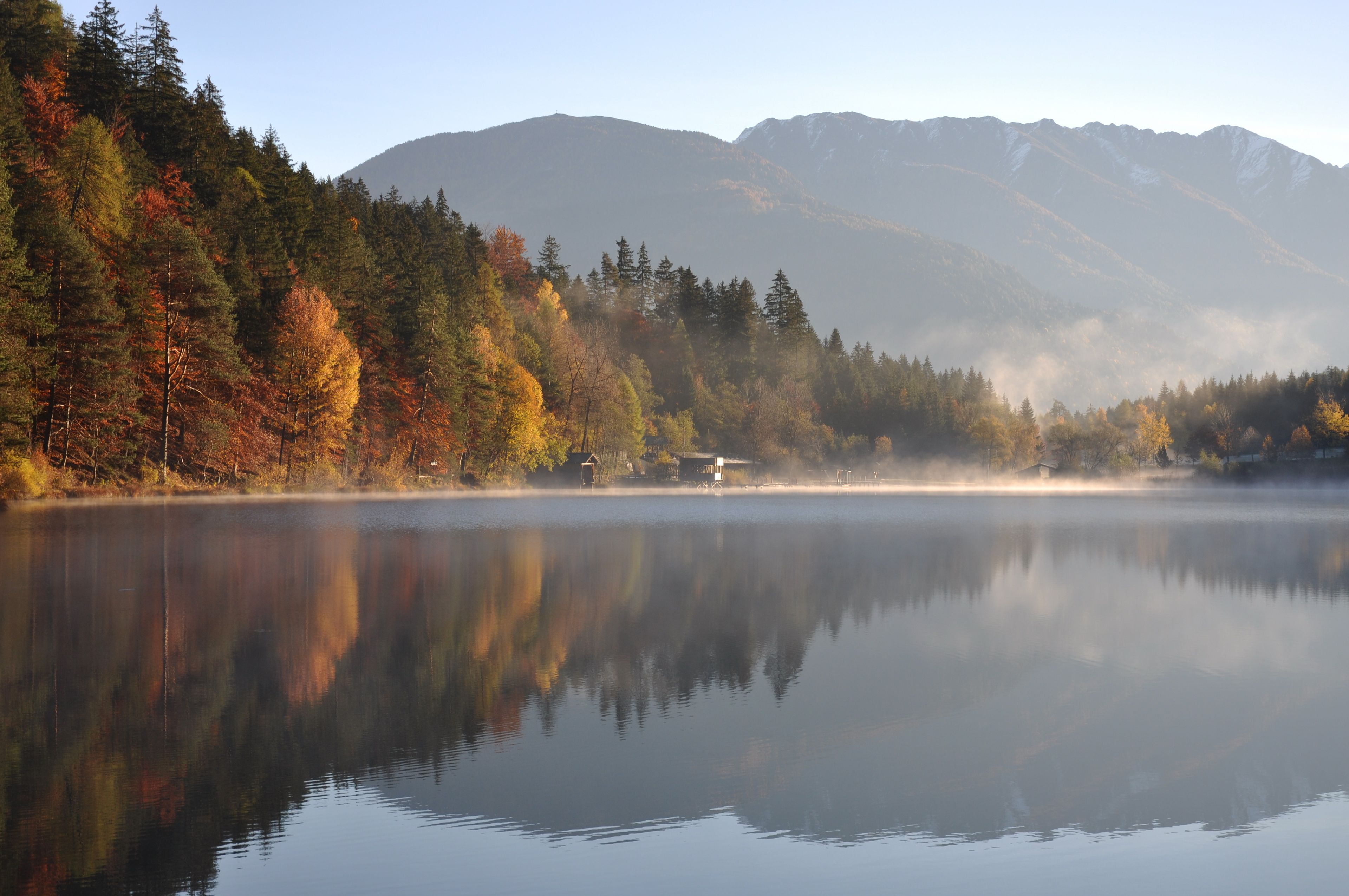 Ein ruhiger See mit Spiegelungen von Bäumen und Bergen im Hintergrund.