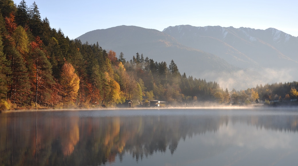 Ein ruhiger See mit Spiegelungen von Bäumen und Bergen im Hintergrund.