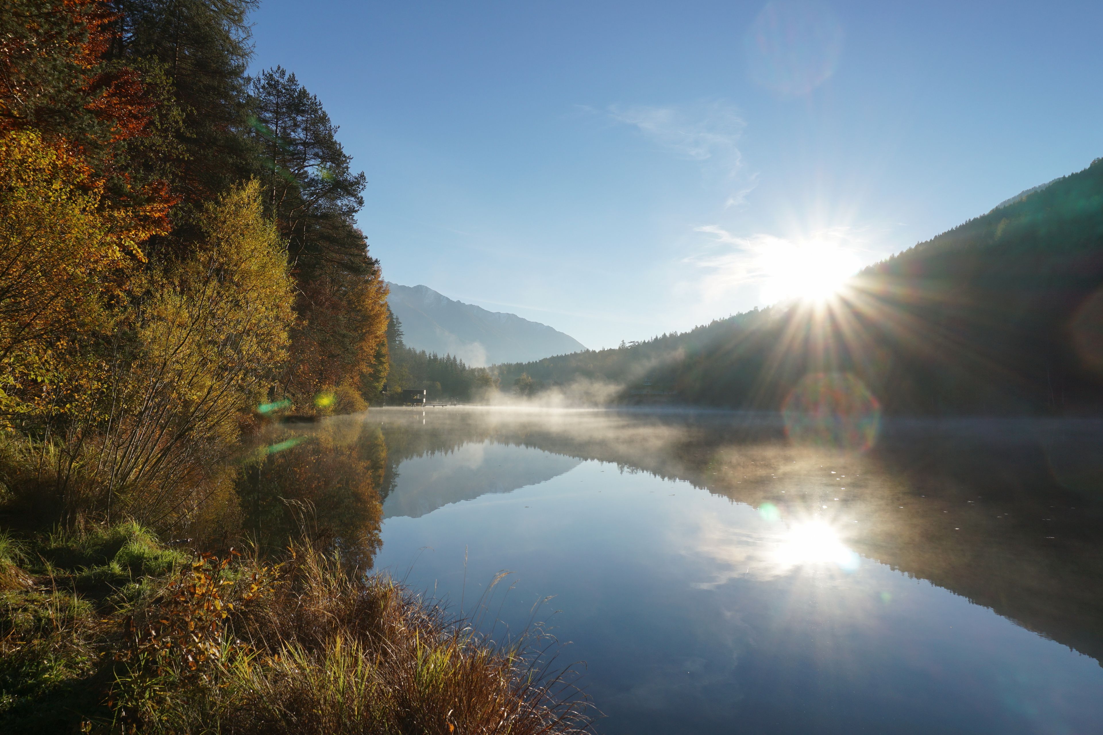 Ein stiller See mit Spiegelungen von Bäumen und Bergen, umgeben von einem Wald im Herbstlaub.