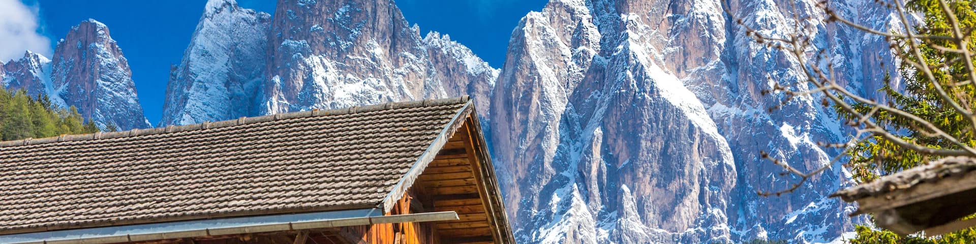 A wooden cabin with a tiled roof, surrounded by snow-covered mountains and a clear blue sky.