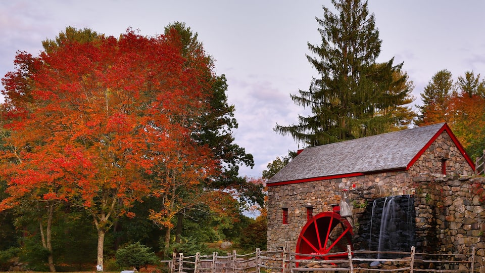 A stone building with a red water wheel and a waterfall, surrounded by autumn-colored trees.