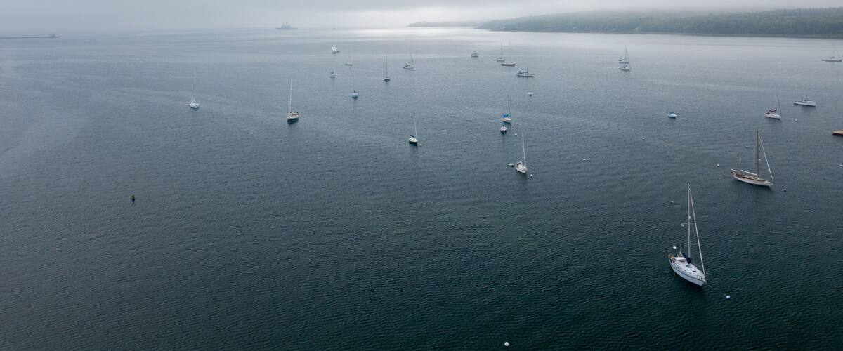 A calm sea with multiple sailboats anchored, a distant ship, and a hazy shoreline.