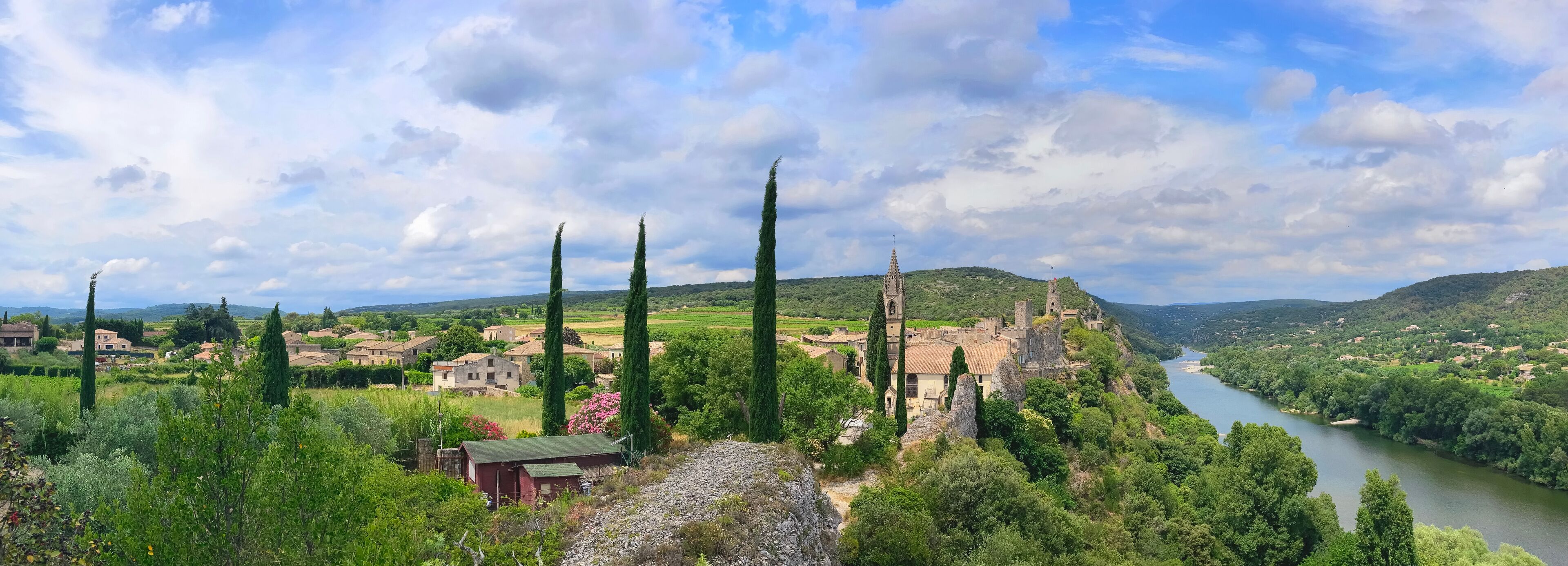 A village with a river, tall cypress trees, and a castle-like structure.