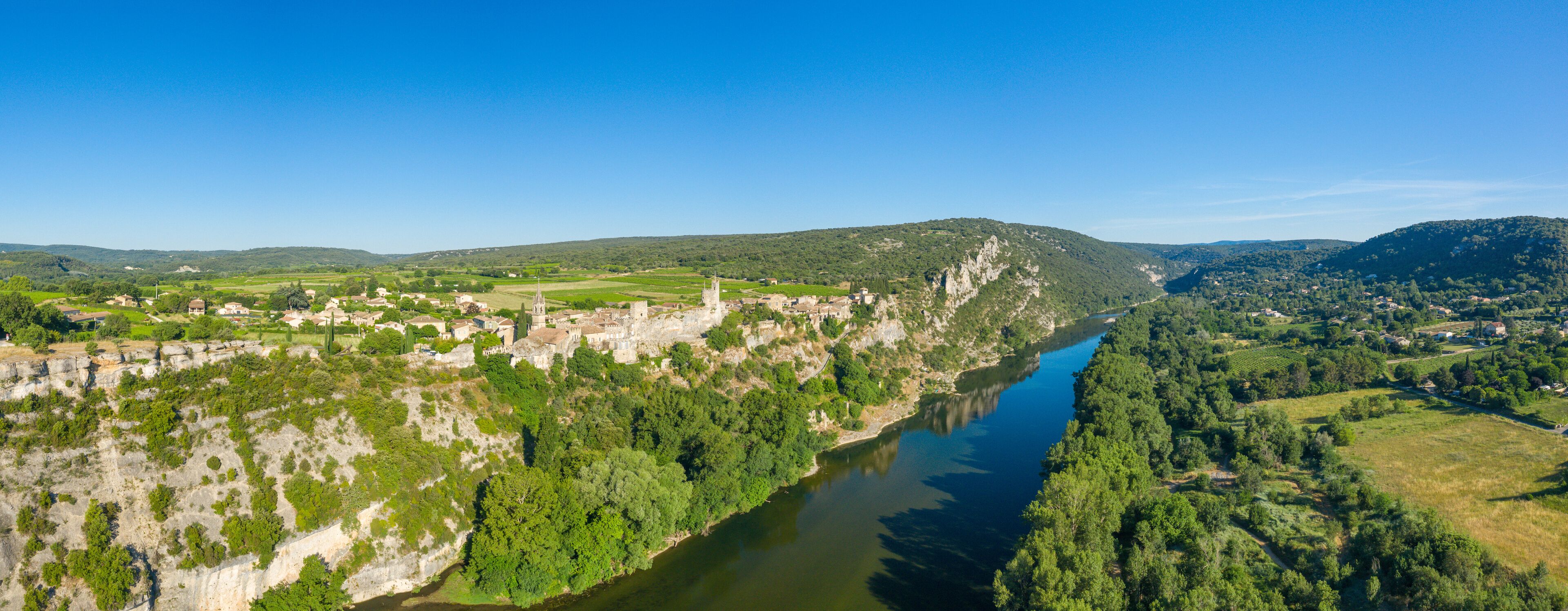 Expansive aerial panorama showing the village of Aigueze perched on limestone cliffs above the winding Ardeche river, surrounded by lush green forests, fields, and distant hills under a clear blue sky