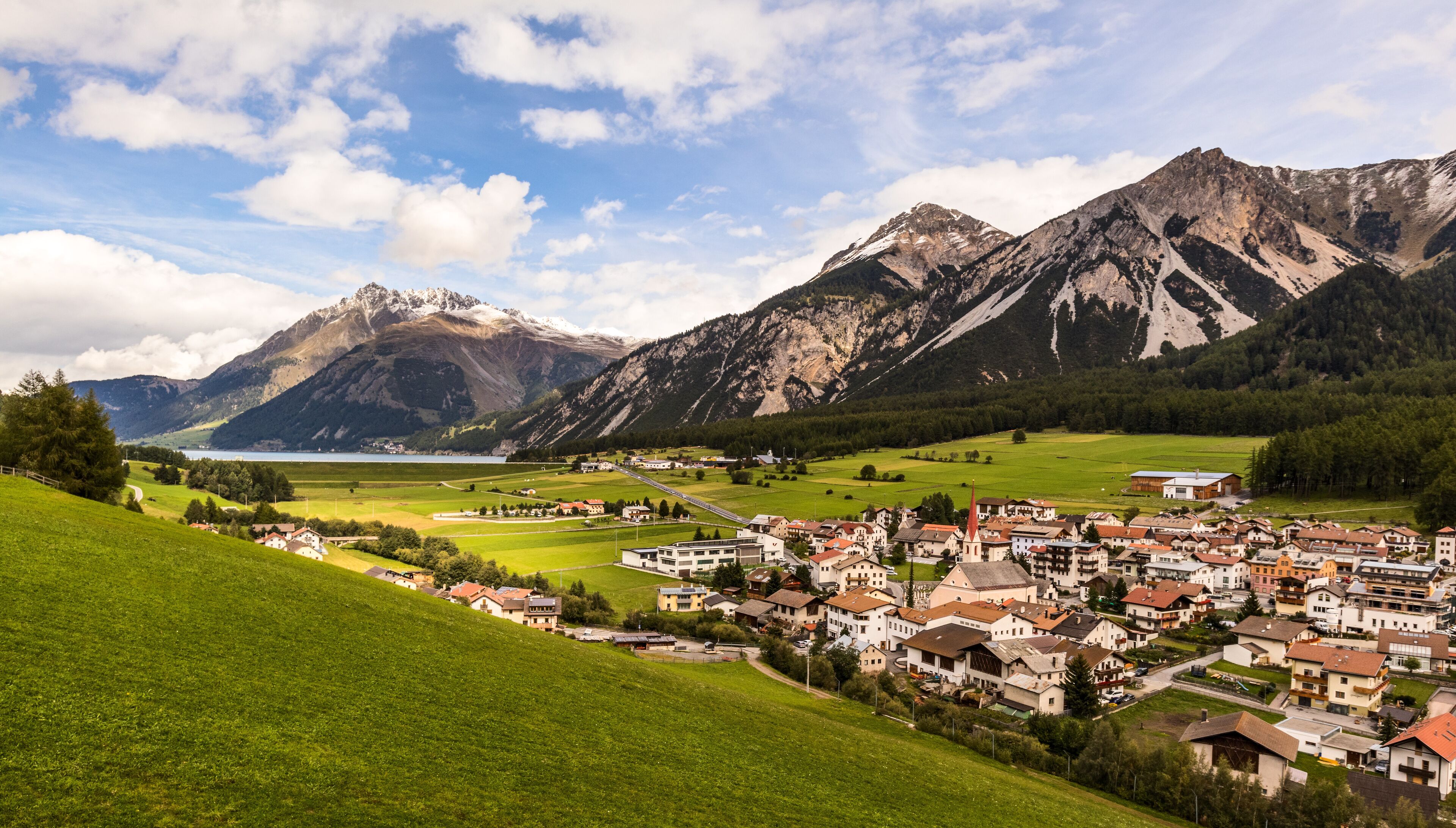 A mountainous landscape with a town nestled in the valley, a lake, and snow-capped peaks in the background.