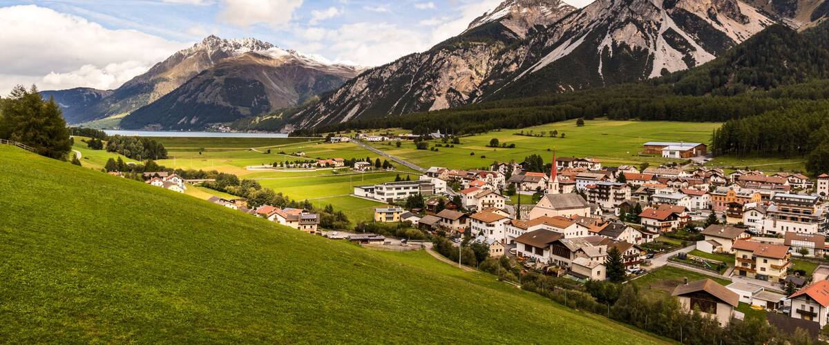 A mountainous landscape with a town nestled in the valley, a lake, and snow-capped peaks in the background.