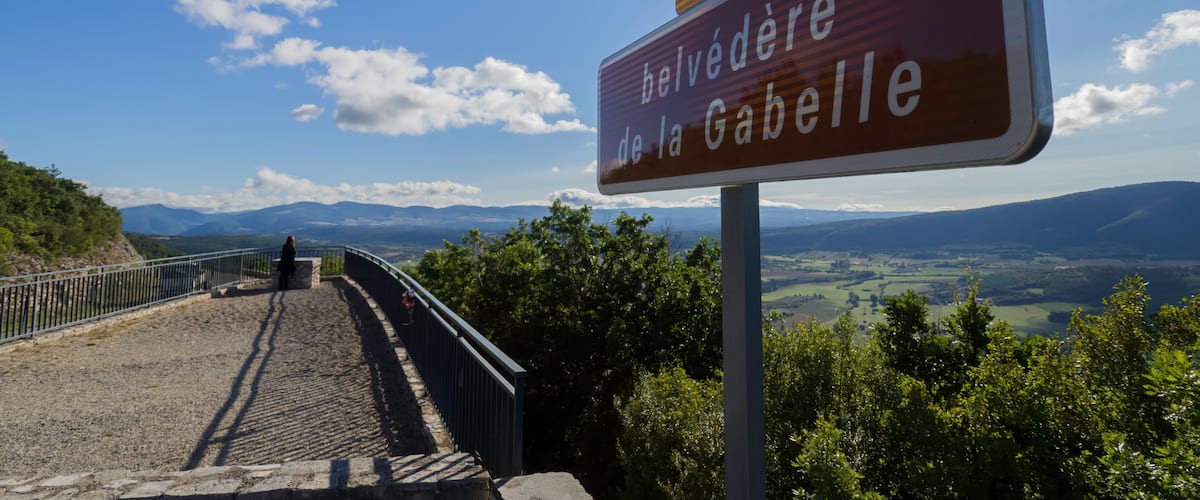 A sign with 'Belvedere de la Gabelle' and 'D1' on a scenic overlook with a view of mountains and a paved path.