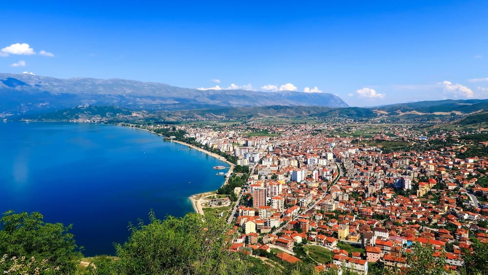 City buildings with red roofs at the shore of a lake with mountains in the background. View of Pogradec city and Ohrid lake, Pogradec, Albania