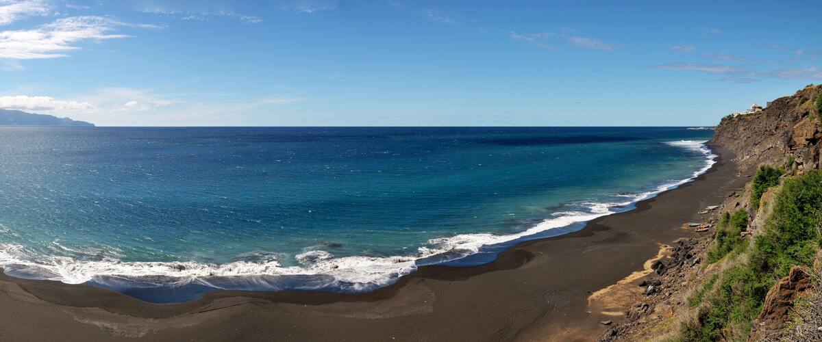 A beach with dark sand, clear blue water, and a rocky cliff.