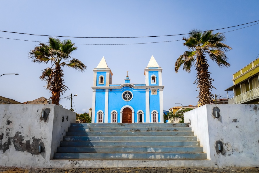 A blue church with two towers, palm trees, and a set of stairs leading to the entrance.