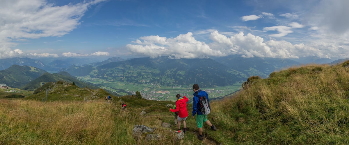 Hikers on a grassy mountain trail with a view of a valley and distant mountains under a partly cloudy sky.