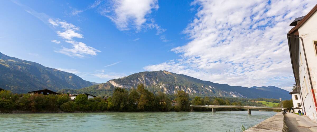 A river with a bridge, houses along the riverbank, and mountains in the background.