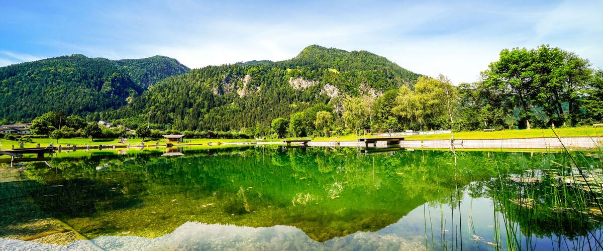 A calm lake with clear reflections, surrounded by lush green hills and a few buildings in the distance.