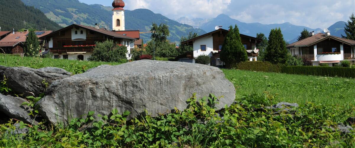 A village with traditional houses, a church with a clock tower, and mountains in the background.