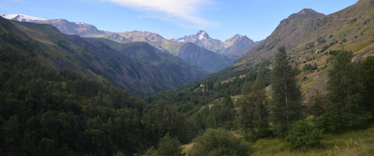 A mountainous landscape with a valley, dense forests, and snow-capped peaks under a partly cloudy sky.