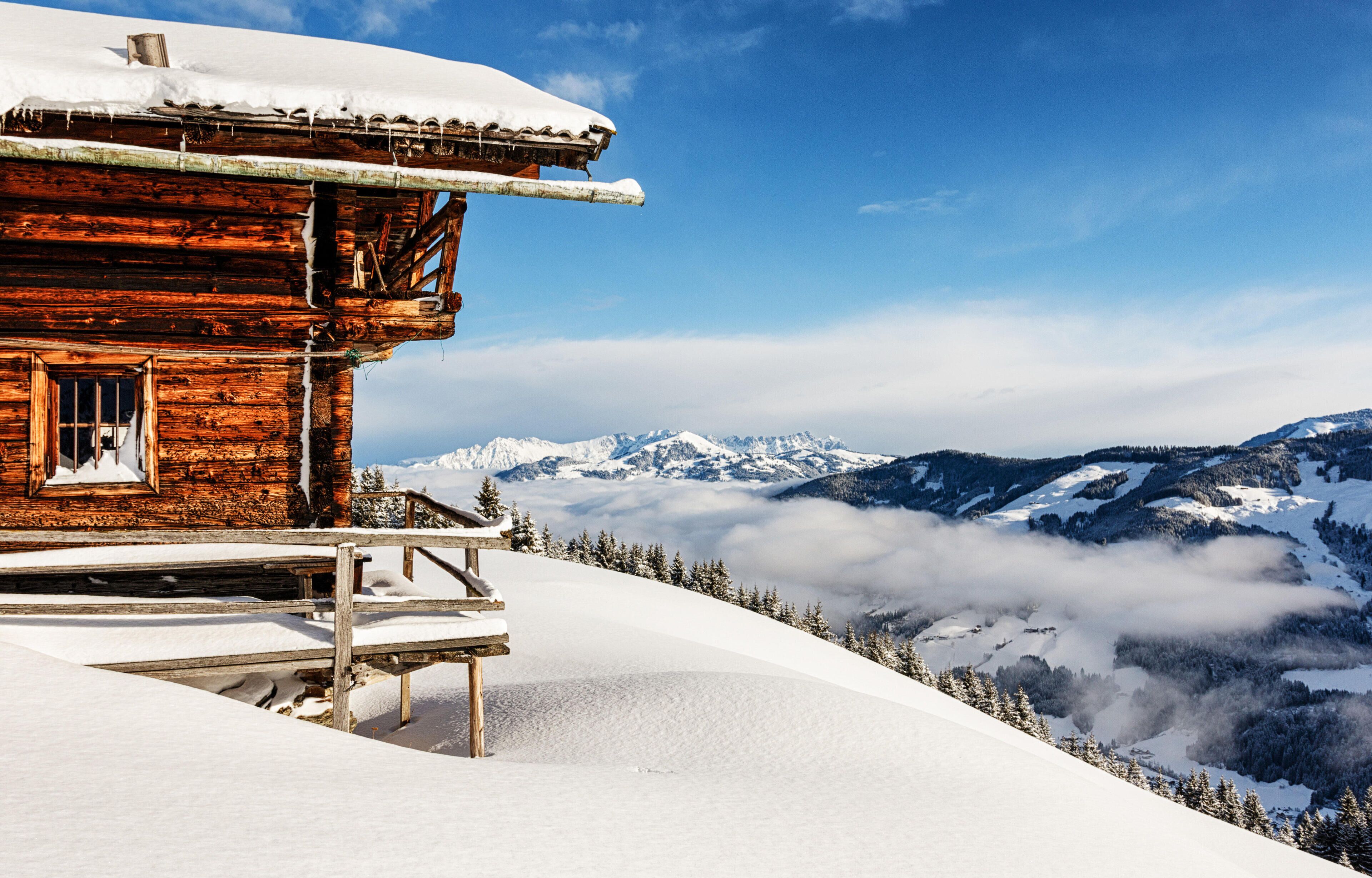A wooden cabin with a snow-covered roof, surrounded by snow-covered mountains and trees.