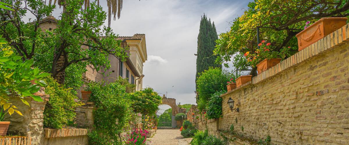 A cobblestone alley flanked by stone walls and lush greenery, leading to a historic building with a tower.