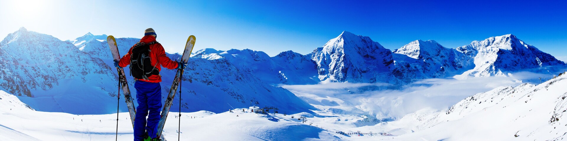 A skier on a snowy mountain with a clear blue sky and snow-covered peaks in the background.