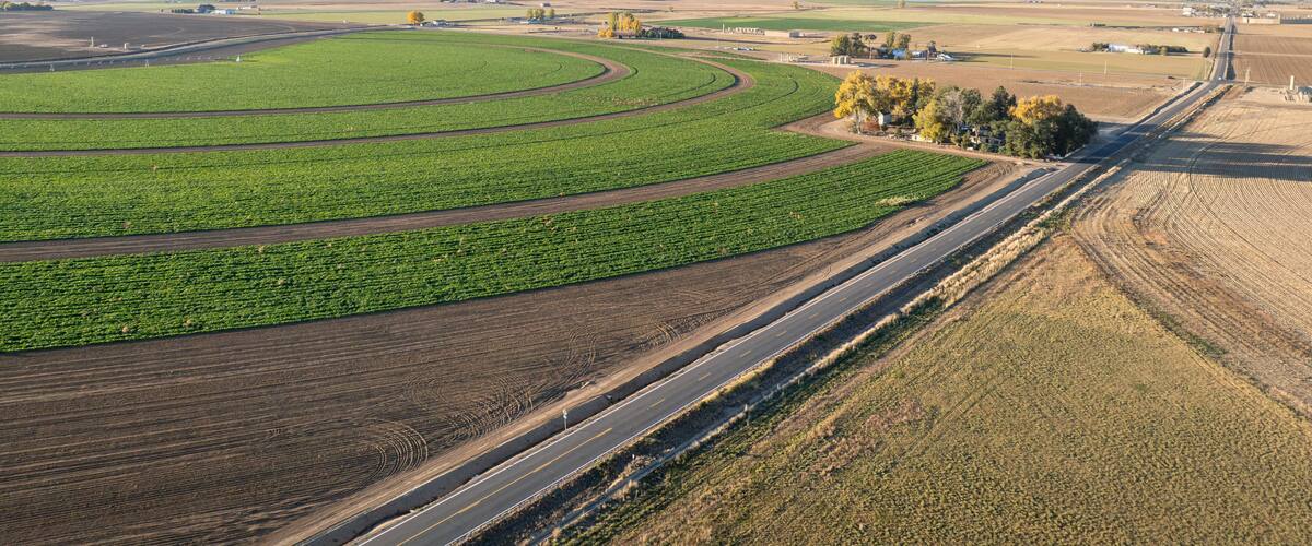 A vast agricultural landscape with green fields, a curved road, and distant buildings.
