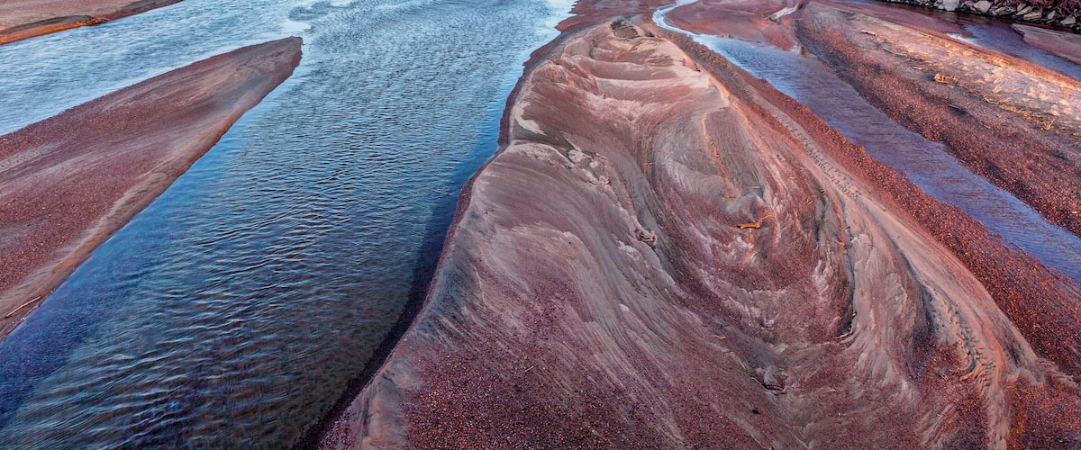 A river with reddish-brown sediment banks and a clear blue waterway.