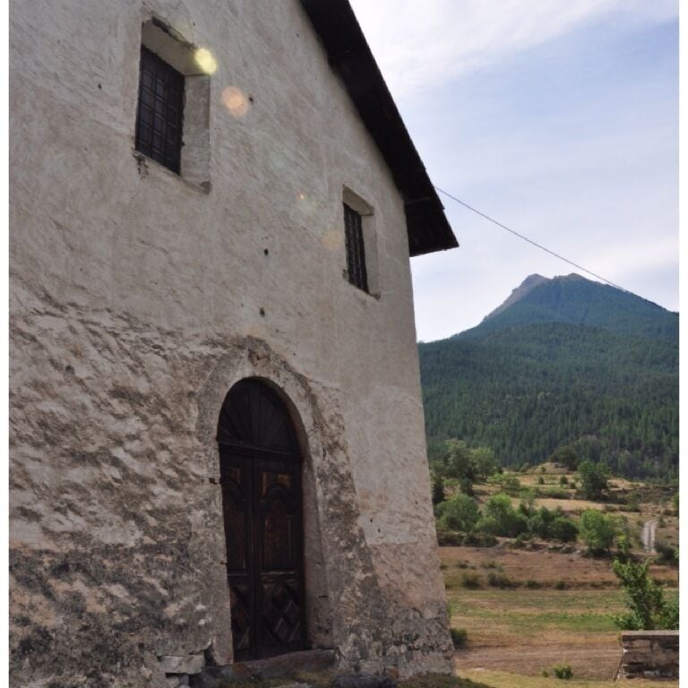 A rustic building with a wooden door and window, set against a backdrop of a mountain and a clear sky.