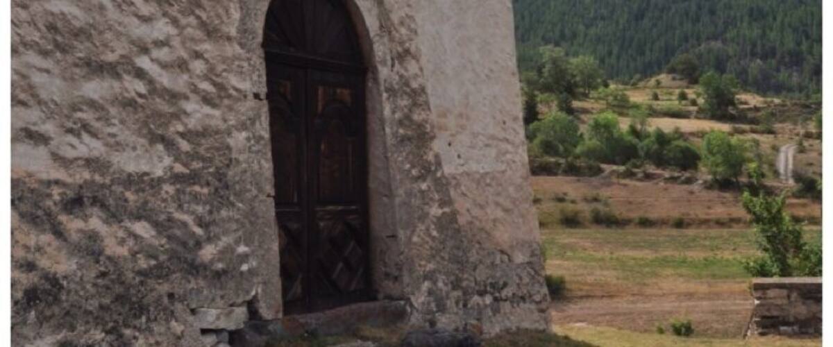A rustic building with a wooden door and window, set against a backdrop of a mountain and a clear sky.