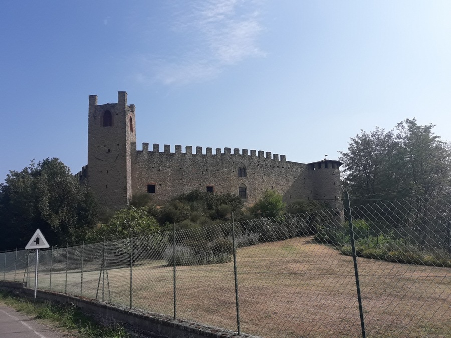 Un castello storico in pietra con un'alta torre e merlature, circondato da una recinzione in catena e vegetazione.