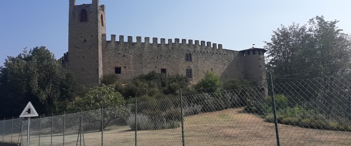 A historic stone castle with a tall tower and battlements, surrounded by a chain-link fence and greenery.