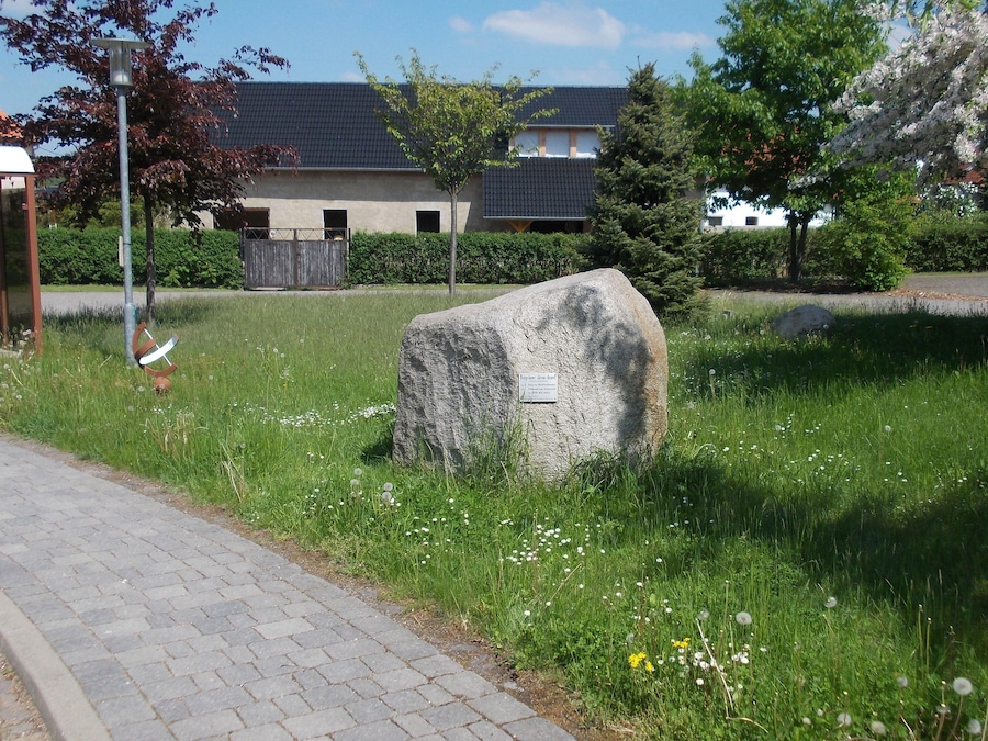 Monumento de piedra en un área con césped, con un camino pavimentado cerca, rodeado de árboles y un edificio con tejado de tejas.