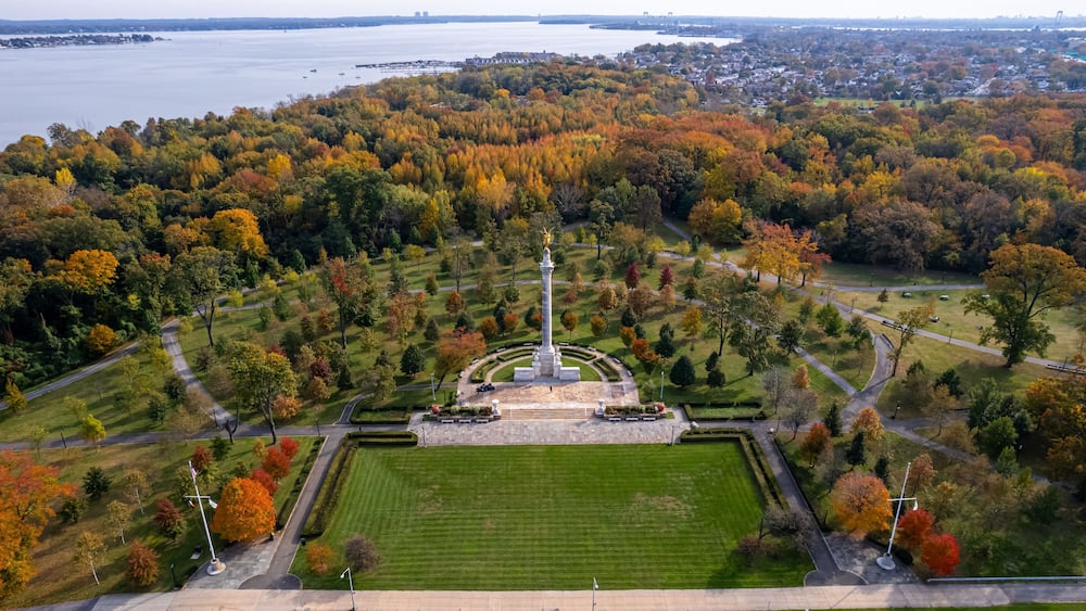 An aerial view of a park with a central monument, surrounded by autumn-colored trees and a body of water in the background.